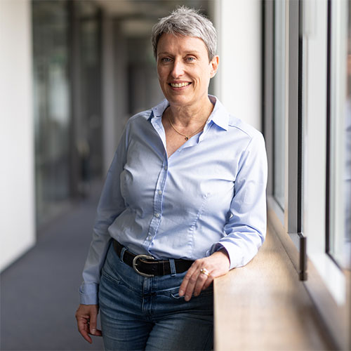 Antje Heide-Picht Antje Heide-Picht, eine Frau mit hellblauer Bluse, Jeanshose und kurzen grauen Haaren, steht in einem Büroflur und lacht in die Kamera.