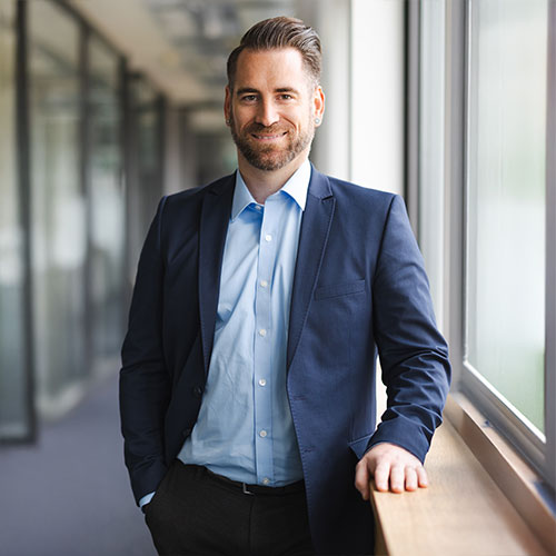 Marco Weinmann Marco Weinmann, ein Mann in dunkelblauen Anzug und hellblauen Hemd steh in einem Büroflur und lacht in die Kamera.